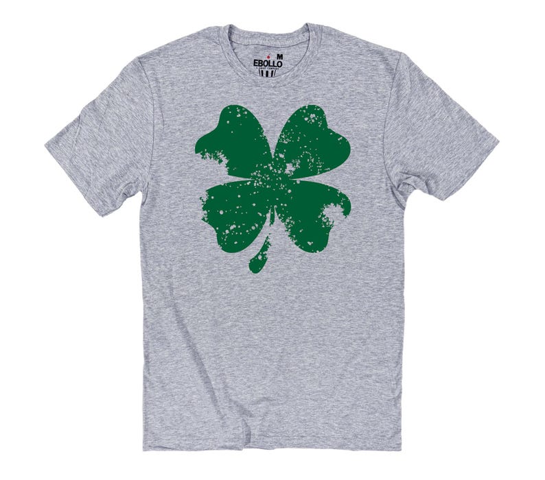 May include: A heather gray t-shirt featuring a large, distressed green shamrock design. The shamrock has a textured, worn appearance. The shirt has short sleeves and a crew neck. The brand name "EBOLLO" is printed on the collar.