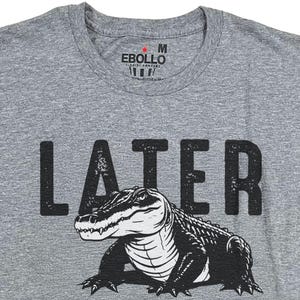 Op de afbeelding: Grijs T-shirt met een zwarte alligator-afbeelding en het woord "LATER" in het zwart gedrukt.