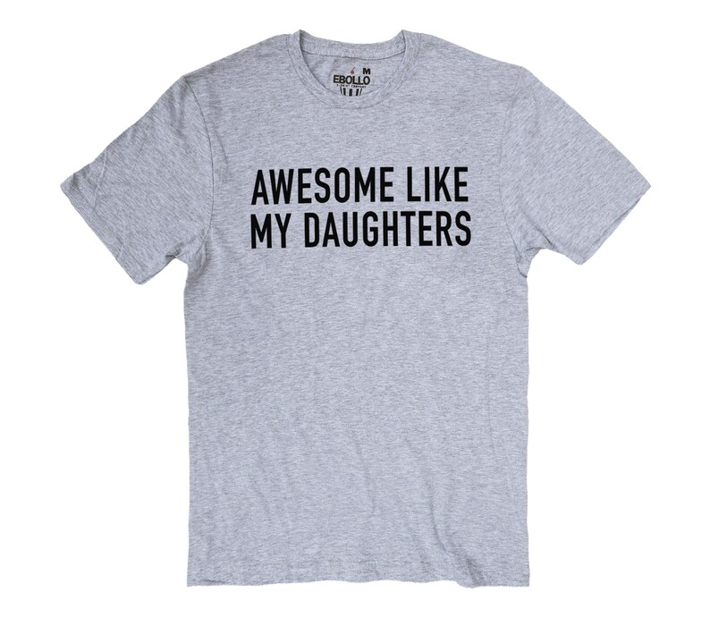 May include: A grey t-shirt featuring the black text "AWESOME LIKE MY DAUGHTERS". The short-sleeved top has a crew neck and the brand name "EBULLO" printed on the upper back. A casual, everyday garment.