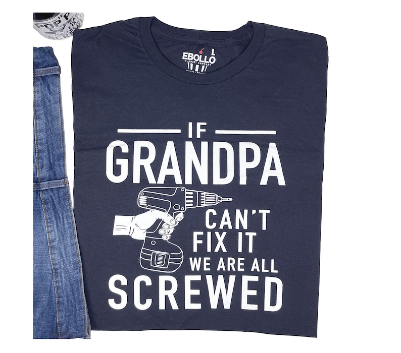 Puede incluir: Camiseta azul marino con el texto blanco "IF GRANDPA CAN'T FIX IT WE ARE ALL SCREWED" y un dibujo de un taladro. La camiseta se muestra junto a un peto vaquero.
