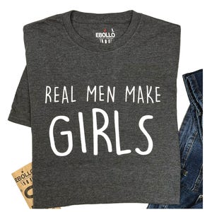 May include: A dark gray t-shirt with the text "REAL MEN MAKE GIRLS" in white. The shirt has a crew neck and a tag that says "EBOLIO". A pair of blue jeans is visible on the right side of the shirt.