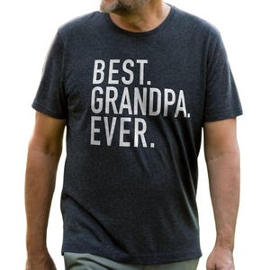 May include: Dark gray t-shirt with the text "BEST. GRANDPA. EVER." in white block letters. The shirt is a crew neck style and appears to be made of a soft, comfortable material. The image shows the shirt on a person.