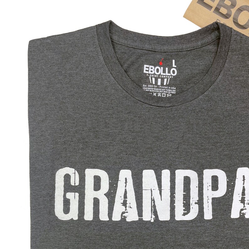 May include: A dark gray t-shirt with the word "GRANDPA" printed in white on the front. The shirt has a crew neck and short sleeves. The t-shirt is made by the Ebollo T-Shirt Company.