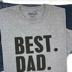 Puede incluir: Una camiseta gris jaspeada talla XL con las palabras "BEST. DAD." impresas en letras negras grandes. La camiseta est&aacute; doblada y colocada sobre unos vaqueros azules. La etiqueta de la marca "Ebollo" es visible.