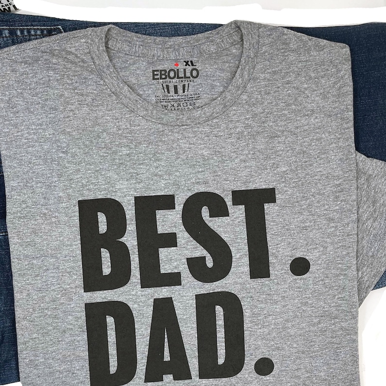Puede incluir: Una camiseta gris jaspeada talla XL con las palabras "BEST. DAD." impresas en letras negras grandes. La camiseta est&aacute; doblada y colocada sobre unos vaqueros azules. La etiqueta de la marca "Ebollo" es visible.