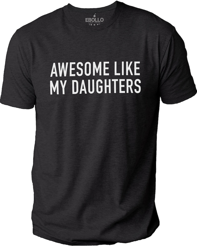 May include: Dark grey t-shirt featuring the white text "AWESOME LIKE MY DAUGHTERS". The shirt has a crew neck and short sleeves. The brand logo is visible at the top.