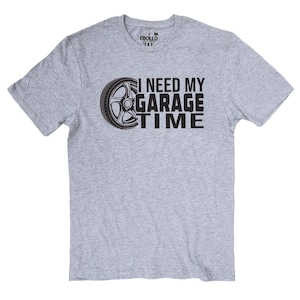 May include: A light gray t-shirt with the text "I NEED MY GARAGE TIME" in black, accompanied by a graphic of a car wheel. The shirt is a medium size and has a crew neck.