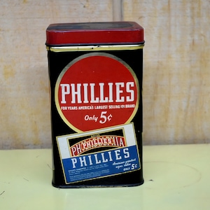 Könnte beinhalten: Eine rechteckige schwarze Metalldose mit rotem Deckel. Die Vorderseite zeigt einen großen roten Kreis mit dem weißen Text PHILLIES und FOR YEARS AMERICA'S LARGEST SELLING 10C BRAND sowie Only 5c. Darunter befindet sich ein rechteckiges Etikett mit PHILLIES in Weiß und Blau.