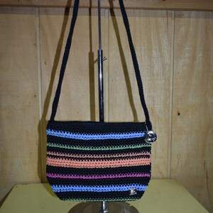 May include: A black crocheted handbag featuring horizontal stripes in light blue, lime green, peach, and magenta. The bag has a single black shoulder strap, a silver-toned circular charm attached to the side, and a small rectangular brand tag.