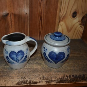 Puede incluir: Un juego de jarra para crema y tarro con tapa de cerámica. Las piezas son de color gris claro con diseños de corazón y hojas azules. La jarra tiene un asa y un pico. El juego está sobre una superficie de madera.