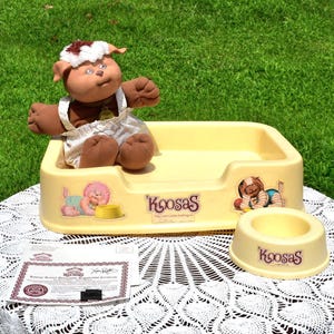 May include: A vintage Koosas doll set, featuring a plush doll with a white bonnet, sitting in a yellow bed. The set includes a matching food bowl and a certificate. The bed and bowl have Koosas branding.