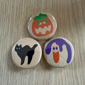 May include: Three circular pinback buttons with a light tan background. One button features an orange jack-o'-lantern with a green stem, one shows a black cat silhouette, and the third displays a white ghost with purple accents and an orange mouth.