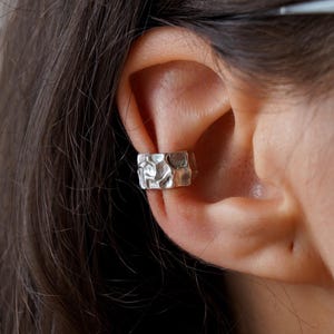 Può includere: Un ear cuff in argento caratterizzato da una fascia larga con superficie testurizzata e martellata. Il metallo presenta una finitura irregolare e craterizzata che crea un aspetto riflettente e disomogeneo sull'intera superficie esterna.