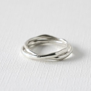 Könnte beinhalten: Ein silberner Ring mit einem einzigartigen, ineinander verschlungenen Design. Der Ring scheint aus einem glänzenden Metall, möglicherweise Sterlingsilber, gefertigt zu sein und weist drei Bänder auf, die sich verdrehen und überlappen. Der Ring ist vor einem weißen Hintergrund platziert.