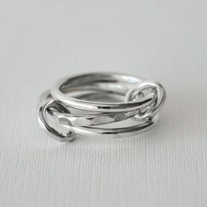Pode incluir: Um anel de prata com um design único. O anel apresenta várias bandas entrelaçadas com um detalhe em forma de nó. O metal tem um acabamento polido, refletindo a luz. O anel é exibido em uma superfície branca.