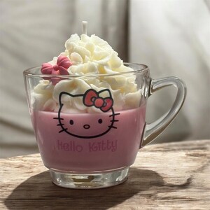Pode incluir: Uma vela em forma de caneca Hello Kitty rosa com chantilly branco e um laço rosa no topo.