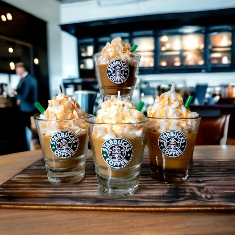 May include: Four Starbucks coffee-themed candles in glass jars with whipped cream and caramel drizzle. Each candle has a green Starbucks logo with the words "Starbucks Coffee" on the front.