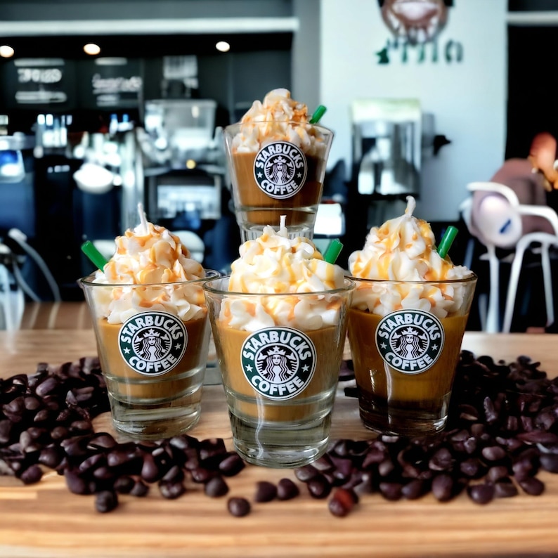 May include: Four Starbucks coffee-themed candles in glass jars with whipped cream and caramel drizzle, surrounded by coffee beans on a wooden surface.