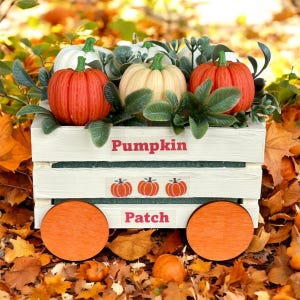 May include: A white wooden wagon filled with pumpkins and greenery, with the words "Pumpkin Patch" on the side. The wagon has orange wheels and is surrounded by autumn leaves. The pumpkins are orange and cream-coloured.