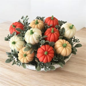 May include: A decorative arrangement of small pumpkins in various colours, including orange, cream, and yellow, nestled among green foliage. The pumpkins are arranged in a white basket, creating a fall-themed centrepiece.