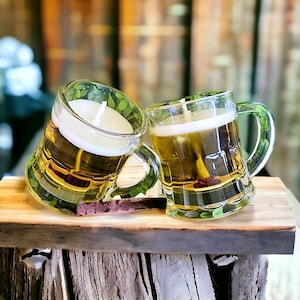 May include: Two clear glass beer mug candles with white wax and wicks. The candles have a yellow-brown liquid wax and are placed on a wooden surface. The mugs have handles and are slightly tilted.