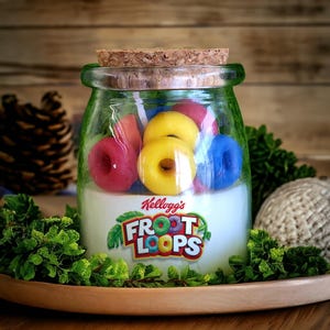 May include: A glass jar candle with a cork lid. The candle is white with the Kellogg's Froot Loops logo on the front. The jar is filled with colorful cereal-shaped candles.