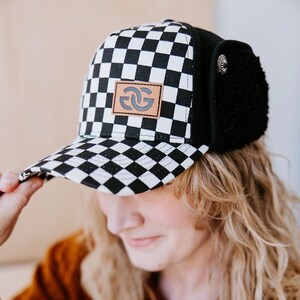 May include: A black and white checkered baseball cap with a leather patch featuring a stylized logo. The cap has a black ear covering with a button detail. The person in the image is wearing a brown shirt.