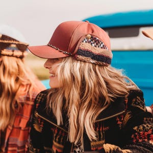 May include: A rust-colored baseball cap with a decorative chain and ear flaps. The ear flaps feature a patterned design with brown, beige, and navy blue accents. The cap is part of a fall fashion collection.