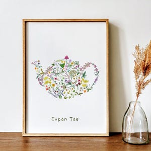 May include: A framed print with a floral teapot design. The teapot shape is filled with a variety of wildflowers in shades of purple, yellow, and pink. The text "Cupan Tae" is printed below the image. The frame is made of wood.