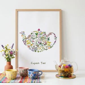 Op de afbeelding: Een ingelijste wandprint met een theepotvorm samengesteld uit kleurrijke wilde bloemen met de tekst Cupan Tae. Daaronder staan drie keramische mokken met bloemmotieven, een vaas met verse bloemen en een glazen theepot met een bloeiende theebloem.