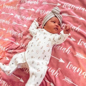 May include: A baby wearing a white floral onesie and a grey turban lies on a pink blanket with the name "Evaniah" repeated in white lettering.