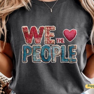 Puede incluir: Camiseta gris oscuro con las palabras "WE THE PEOPLE" en letras rojas y azules desgastadas. Un corazón rojo está encima de la palabra "THE". La camiseta es de estilo casual y cómodo.