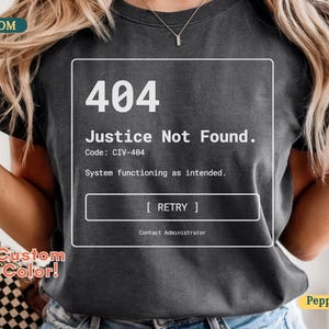 Puede incluir: Camiseta gris oscuro con un gráfico blanco que muestra un mensaje de error 404: "Justice Not Found." El texto incluye "Code: CIV-404" y "System functioning as intended." También es visible un botón "Retry". La camiseta es de color personalizado.