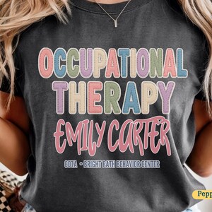 Puede incluir: Una camiseta gris carbón con el texto OCCUPATIONAL THERAPY en letras de bloque multicolores con un efecto de contorno cosido. Debajo, el nombre EMILY CARTER está impreso en letra cursiva rosa, seguido de COTA - BRIGHT PATH BEHAVIOR CENTER.