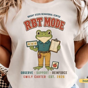 May include: An ivory t-shirt featuring a graphic of a green frog dressed in a blue polo shirt and tan pants. The frog holds a star rating card and a clipboard. Text includes BRIGHT STEPS BEHAVIORAL CENTER, RBT MODE, OBSERVE, SUPPORT, REINFORCE, EMILY CARTER, EST. 2026.