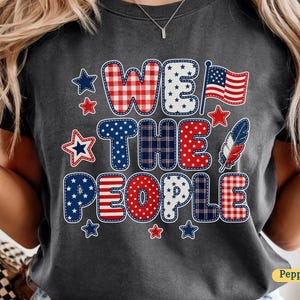 Puede incluir: Camiseta gris oscuro con el texto "WE THE PEOPLE" en un diseño de patchwork. El diseño incorpora elementos rojos, blancos y azules, incluyendo estrellas, una bandera y una pluma, evocando un tema patriótico.
