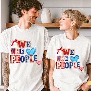 Puede incluir: Camisetas blancas con el texto "WE THE PEOPLE" en rojo, azul y blanco. El diseño incluye un corazón azul y un lazo rojo. Las camisetas son usadas por dos personas.