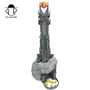 Op de afbeelding: Een donkergrijze toren met een gouden oog bovenop, omringd door een rotsachtige basis. De toren heeft een dobbelsteenrollade onderaan, met daarin verschillende gouden dobbelstenen.