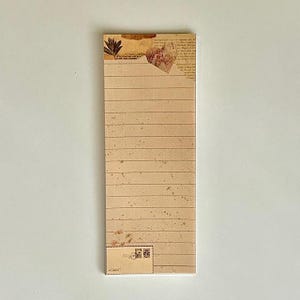 May include: A lined notepad with a vintage aesthetic. The paper is cream colored with brown lines and a subtle speckled pattern. The top of the notepad features a decorative design with a brown leaf and a piece of paper with text.
