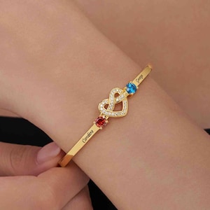 May include: A gold-colored bangle bracelet with a heart-shaped design. The bracelet features a red and blue gemstone, and the names "Caroline" and "George" are engraved on the bracelet.