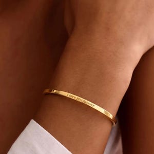 May include: A gold-colored bangle bracelet with the inscription "I love you forever." The bracelet is thin and simple, with a smooth, polished finish. The bracelet is worn on a person's wrist.