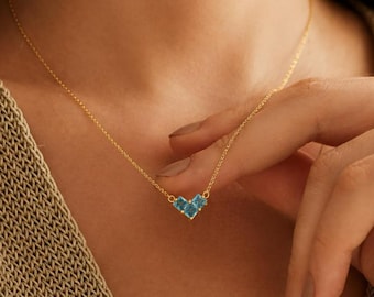May include: A delicate gold necklace featuring a heart-shaped pendant. The pendant is composed of three square, light blue gemstones. The necklace is worn around the neck.