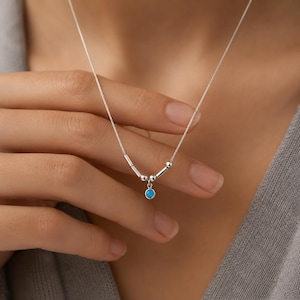 Può includere: Una delicata collana d'argento con un piccolo pendente rotondo turchese. La collana ha una catena sottile con piccole perle d'argento e accenti cilindrici. Il pendente è di un vibrante colore turchese, che aggiunge un tocco di colore al design minimalista.