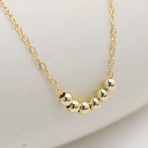 May include: A delicate gold necklace with a series of small, round beads. The beads are evenly spaced and connected by a fine gold chain. The necklace is displayed on a white surface, highlighting its simple elegance.