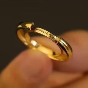 Może przedstawiać: Złoty pierścionek z wygrawerowanym napisem "Michea I & Paige". Pierścionek ma unikalny design z niewielkim otworem. Pierścionek jest wykonany z błyszczącego, odbijającego światło metalu.