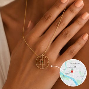 May include: A gold-toned necklace featuring a circular pendant with a cut-out street map design. A small, round green gemstone is set at a specific location on the map. The pendant hangs from a delicate gold-toned chain.