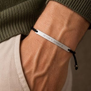 Può includere: Un bracciale con barretta metallica color argento su un sottile cordino nero. La piastra metallica rettangolare è incisa con le coordinate 40.7128° N, 74.0060° W. Il bracciale presenta estremità regolabili con piccole perline metalliche.