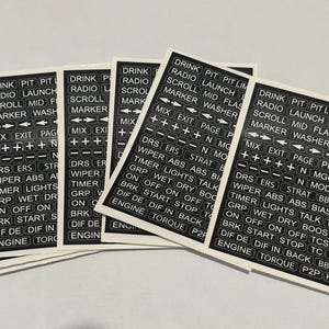 May include: A stack of black and white stickers with various text labels. The labels include words like "DRINK", "RADIO", "LAUNCH", "SCROLL", "MARKER", and "WIPER". The stickers are likely for automotive or racing applications.