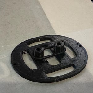 May include: A black, 3D-printed, circular object with an intricate design. It features a central structure with two cylindrical posts connected by a curved bar, and a series of rectangular cutouts around the perimeter. The object is made of a textured material.