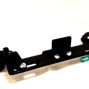 Op de afbeelding: Een zwarte metalen beugel met meerdere bevestigingspunten en een gebogen basis. De beugel heeft een rechthoekig ontwerp met verschillende zilveren schroeven en een klein, lichtblauw steundeel.